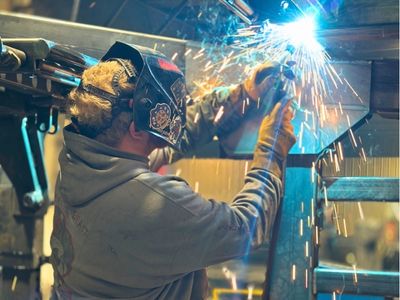 Rows of stacked rectangular and square American-sourced steel tubes and beams stretch into the background of a manufacturing facility, with a person visible in the distance.