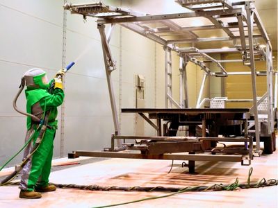 Rows of stacked rectangular and square American-sourced steel tubes and beams stretch into the background of a manufacturing facility, with a person visible in the distance.