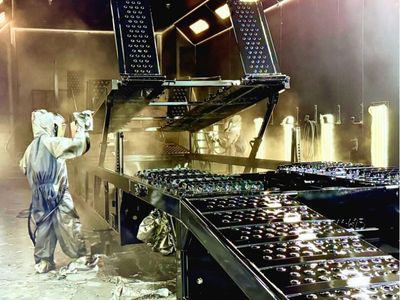 Rows of stacked rectangular and square American-sourced steel tubes and beams stretch into the background of a manufacturing facility, with a person visible in the distance.