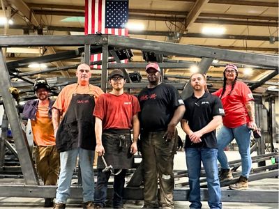 Rows of stacked rectangular and square American-sourced steel tubes and beams stretch into the background of a manufacturing facility, with a person visible in the distance.
