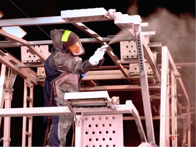 Rows of stacked rectangular and square American-sourced steel tubes and beams stretch into the background of a manufacturing facility, with a person visible in the distance.